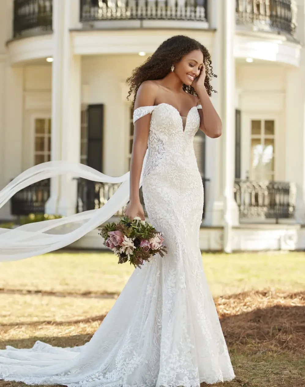 A woman in a white off-the-shoulder wedding dress stands outdoors, holding a bouquet of flowers while smiling. She has curly hair and is standing in front of a building with large columns.