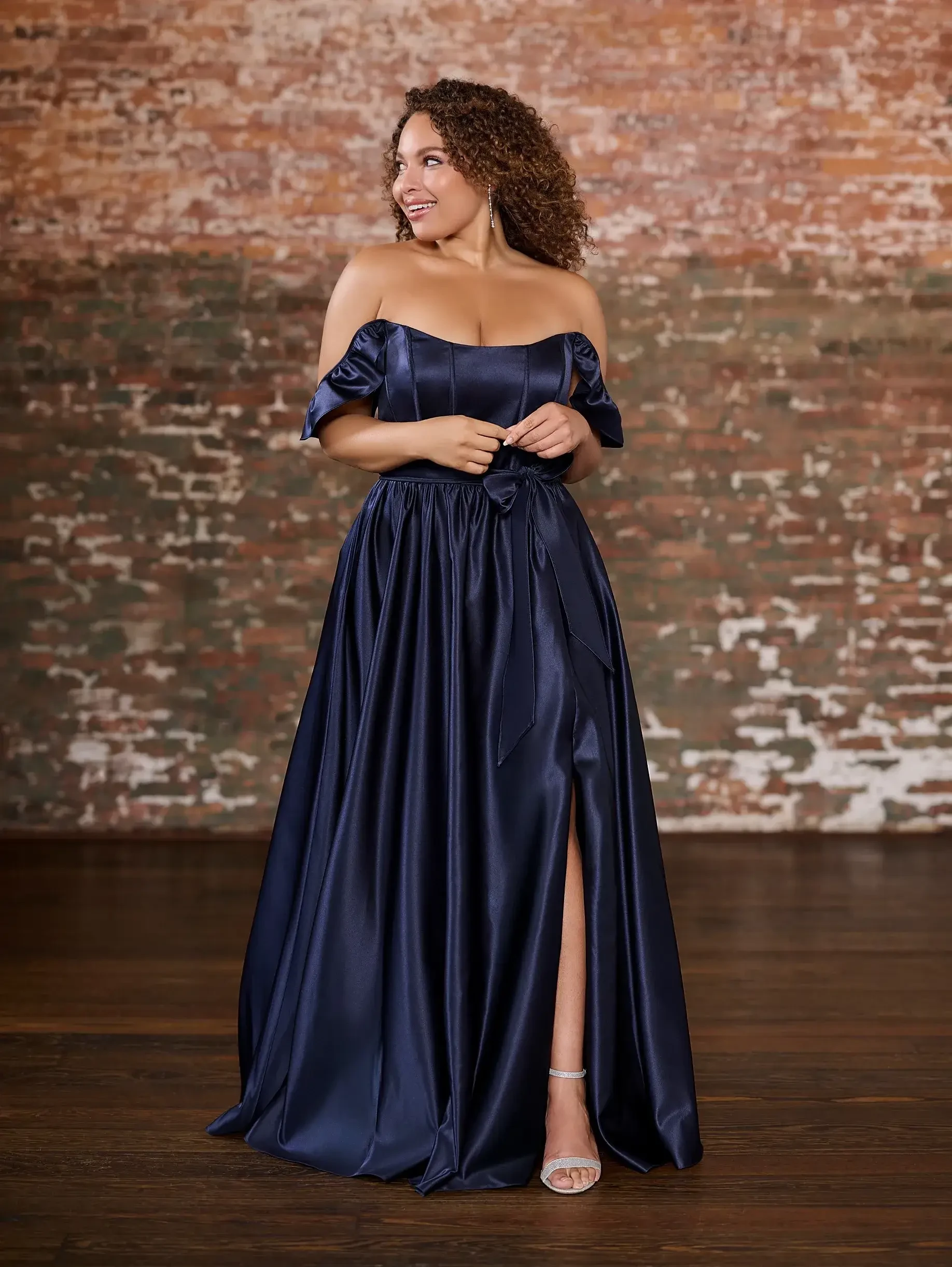 A woman stands gracefully in a navy off-shoulder gown, featuring a high slit and flowing satin fabric. She appears joyful against a rustic brick wall.