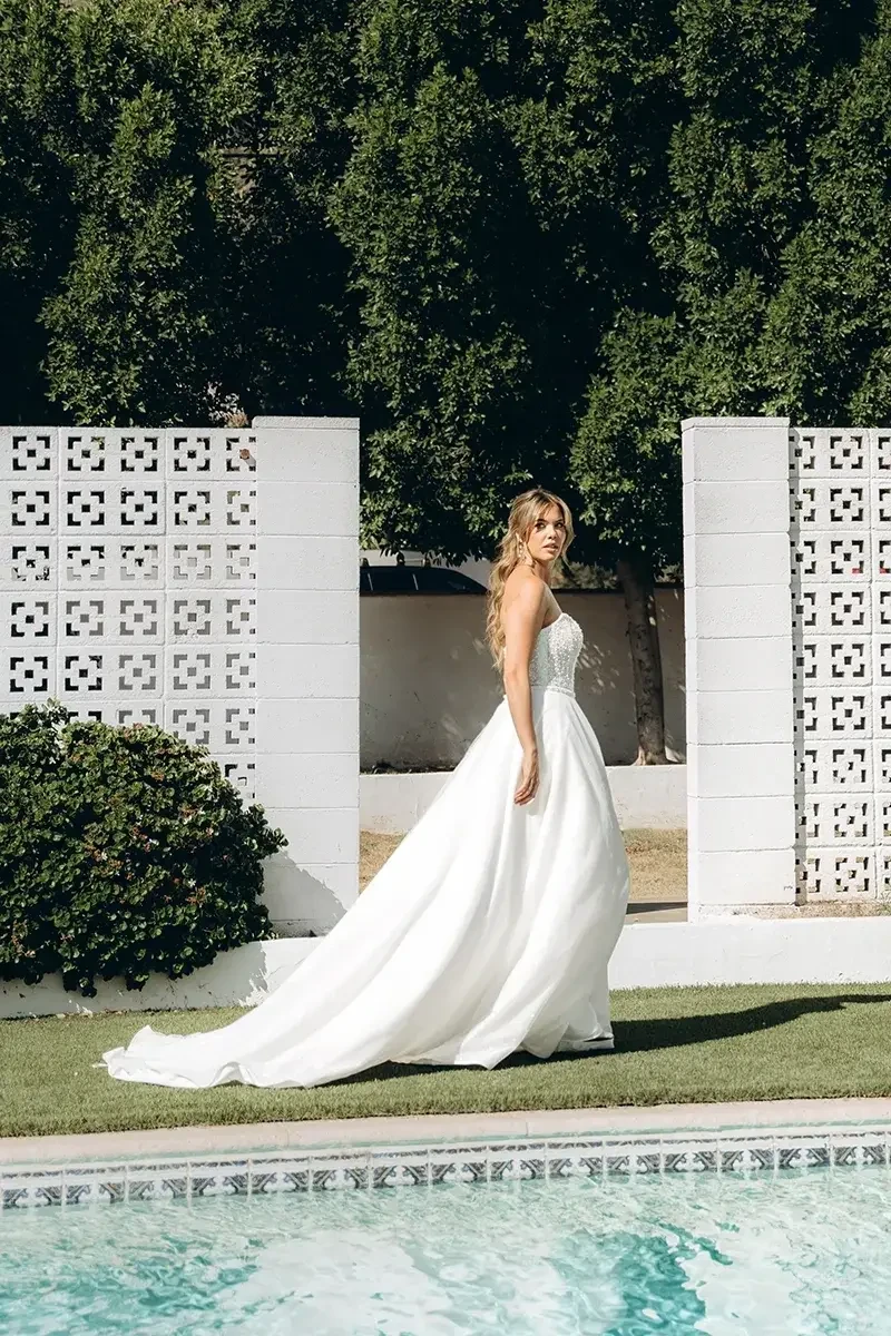Model is wearing a strapless A-line gown with a sheer corset bodice, basque waist, glittering lace, and a detachable tulle and organza overskirt.