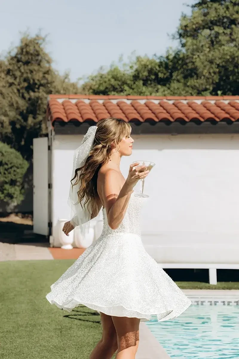 Model is wearing a strapless A-line gown with a sheer corset bodice, basque waist, glittering lace, and a detachable tulle and organza overskirt.