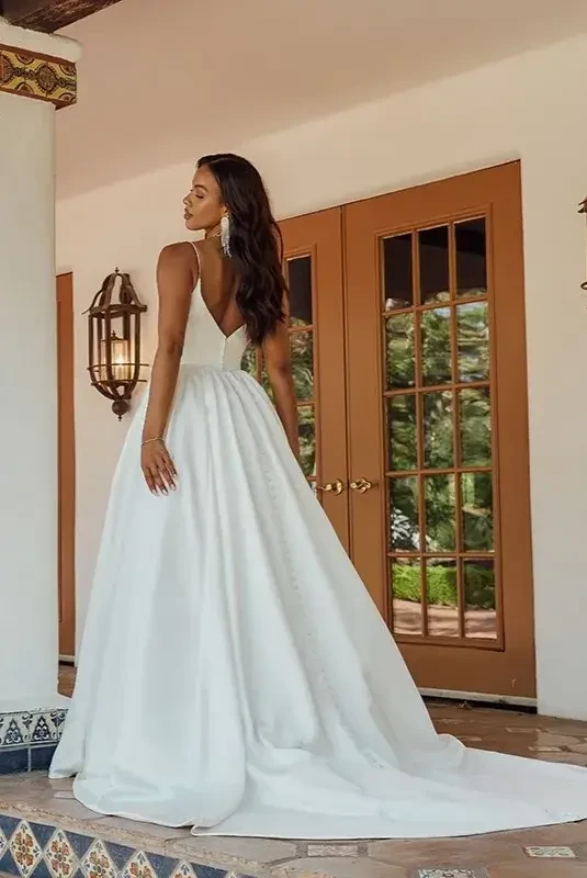 Model is wearing a modern satin ballgown with spaghetti straps, side cutouts, a scooped neckline, V-shaped open back with buttons, and a ruched full skirt with discreet pockets.