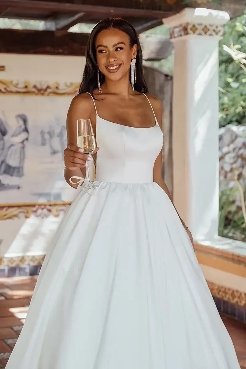 Model is wearing a modern satin ballgown with spaghetti straps, side cutouts, a scooped neckline, V-shaped open back with buttons, and a ruched full skirt with discreet pockets.