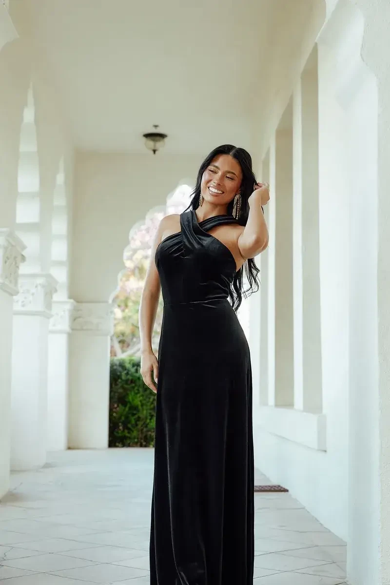 A woman stands smiling in a long black halter dress in a bright, arched corridor. The scene feels elegant and serene, with soft, natural light.