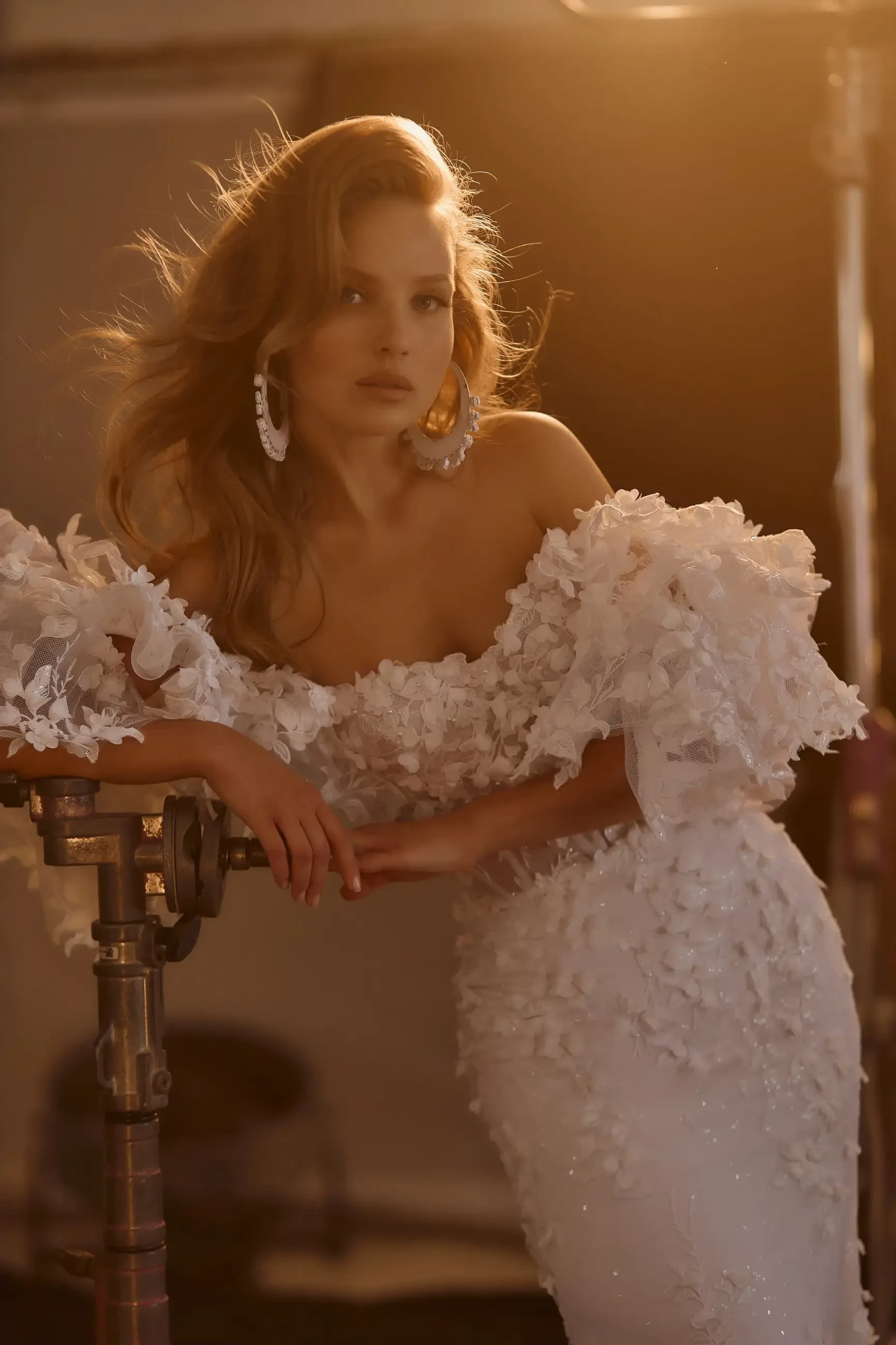 A woman in an elegant white dress with ruffled sleeves poses gracefully, leaning on a prop with soft lighting highlighting her features.
