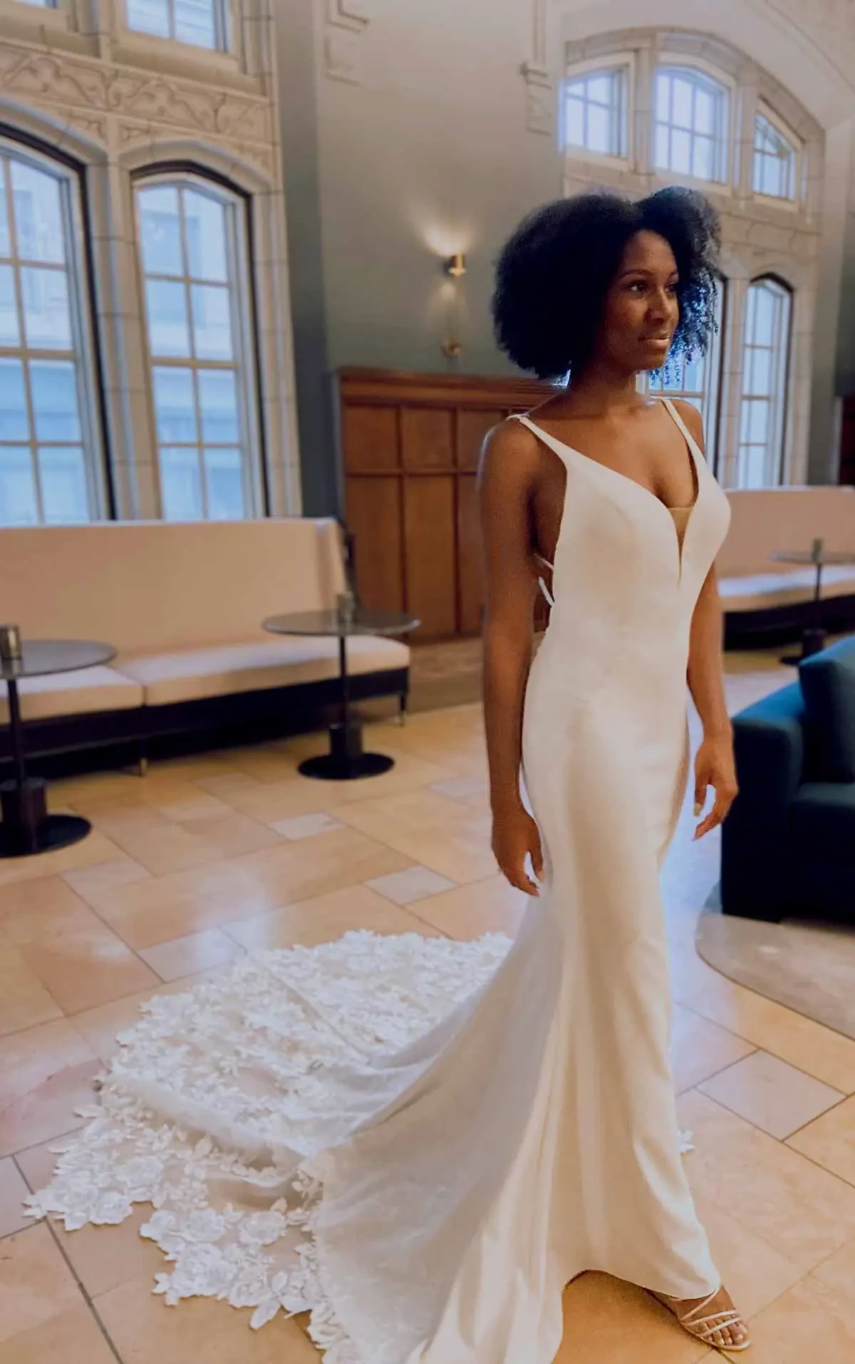 A woman wearing a fitted white wedding dress with a long train, standing in an elegant interior space.