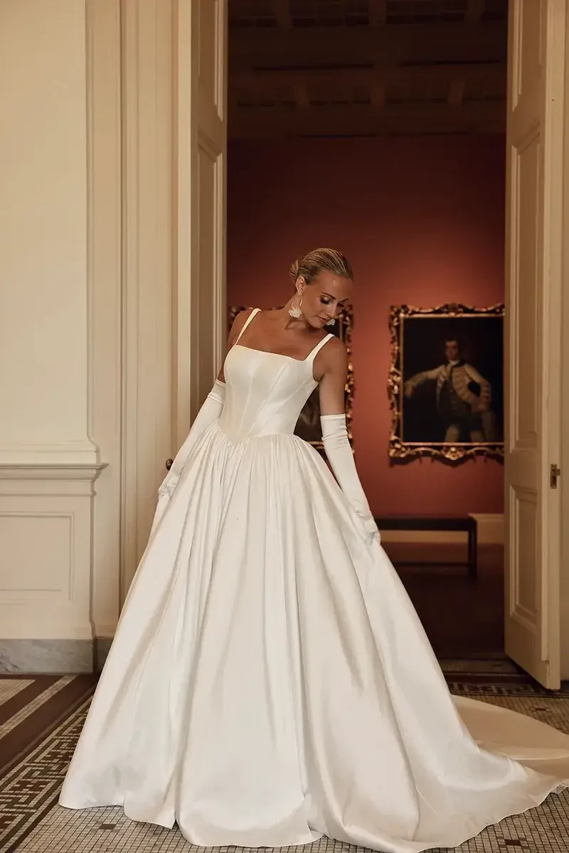 A woman in an elegant white dress with a fitted bodice and full skirt, standing in a classical setting with a painting in the background.