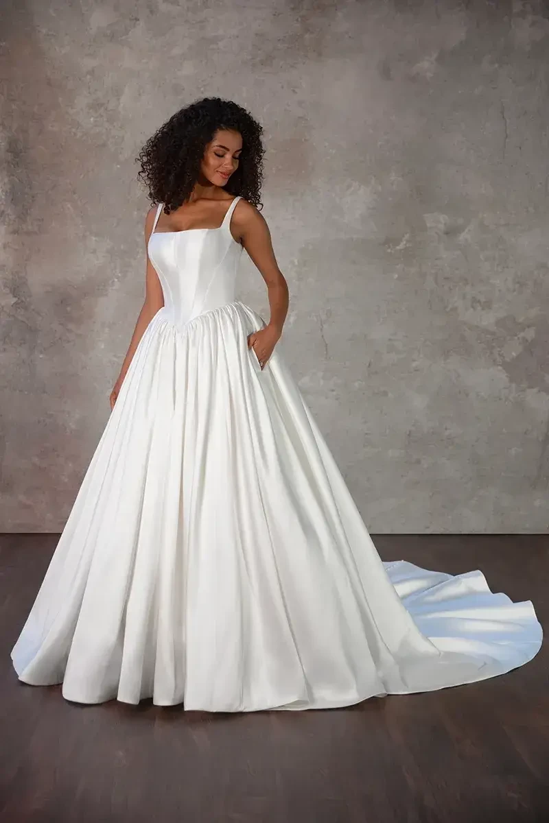 A woman wearing a long, elegant white gown with a full skirt and thin straps, standing against a textured gray background. The dress has pockets and a train flowing behind her. She has curly hair and is looking slightly to the side.