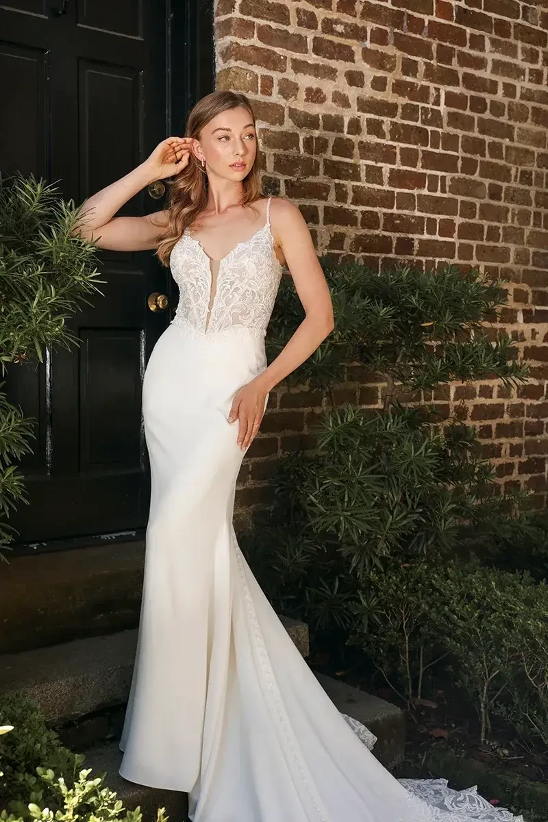 A woman in a fitted white wedding dress with a lace bodice stands outside near a brick wall and greenery.