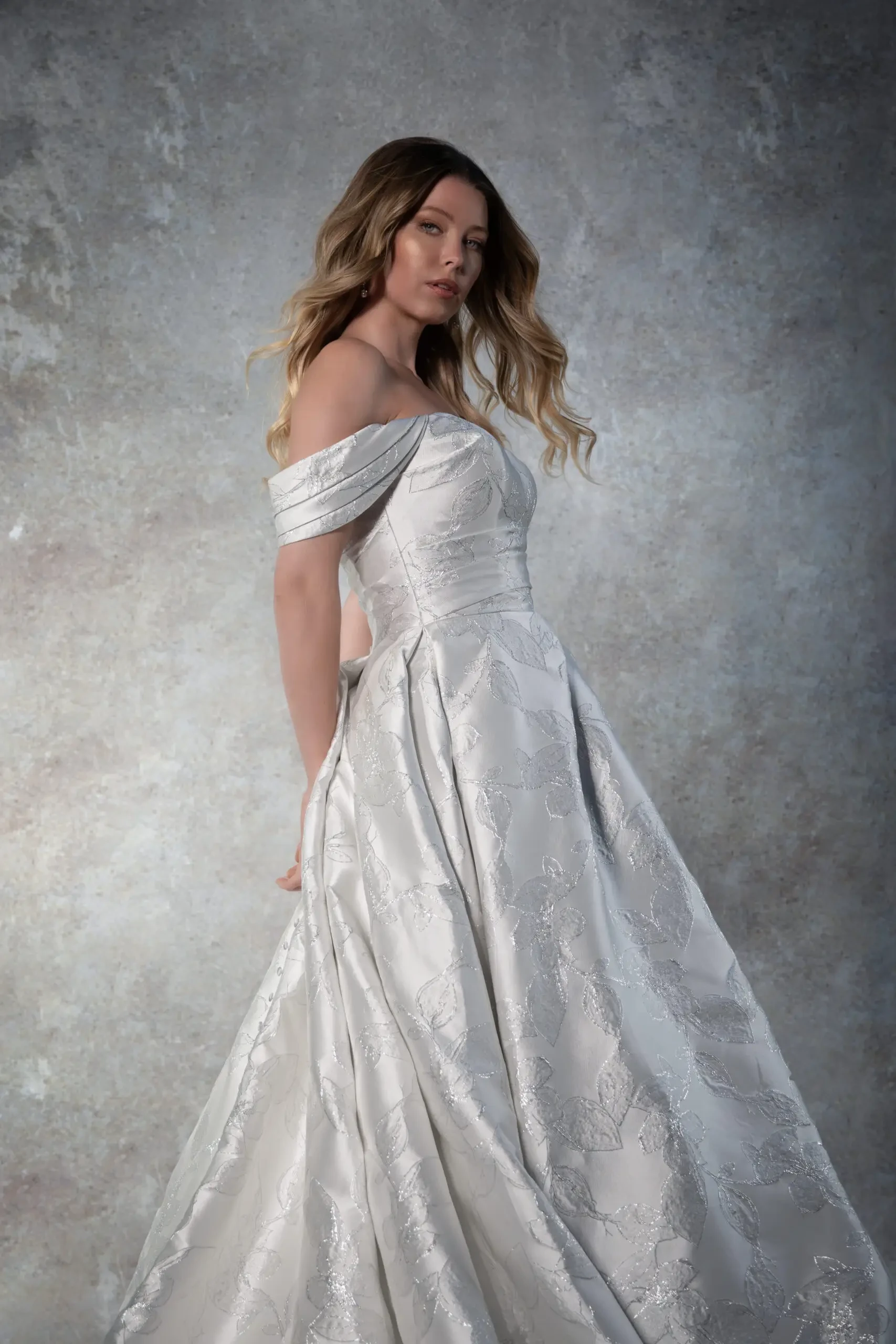 A woman in an elegant, off-shoulder white gown with floral patterns stands against a textured gray background, exuding a poised and sophisticated aura.