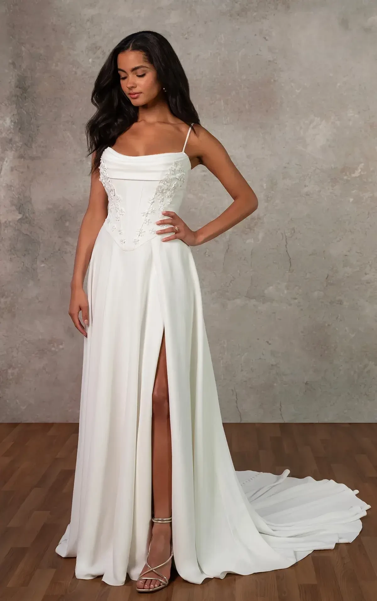 A woman in a white gown with a thigh-high slit and thin straps, standing on wooden flooring against a textured wall.