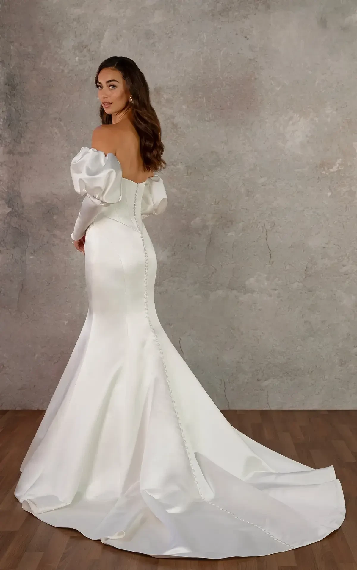 A woman wearing an elegant off-shoulder white gown with a fitted silhouette and a long train, standing sideways against a neutral textured background.