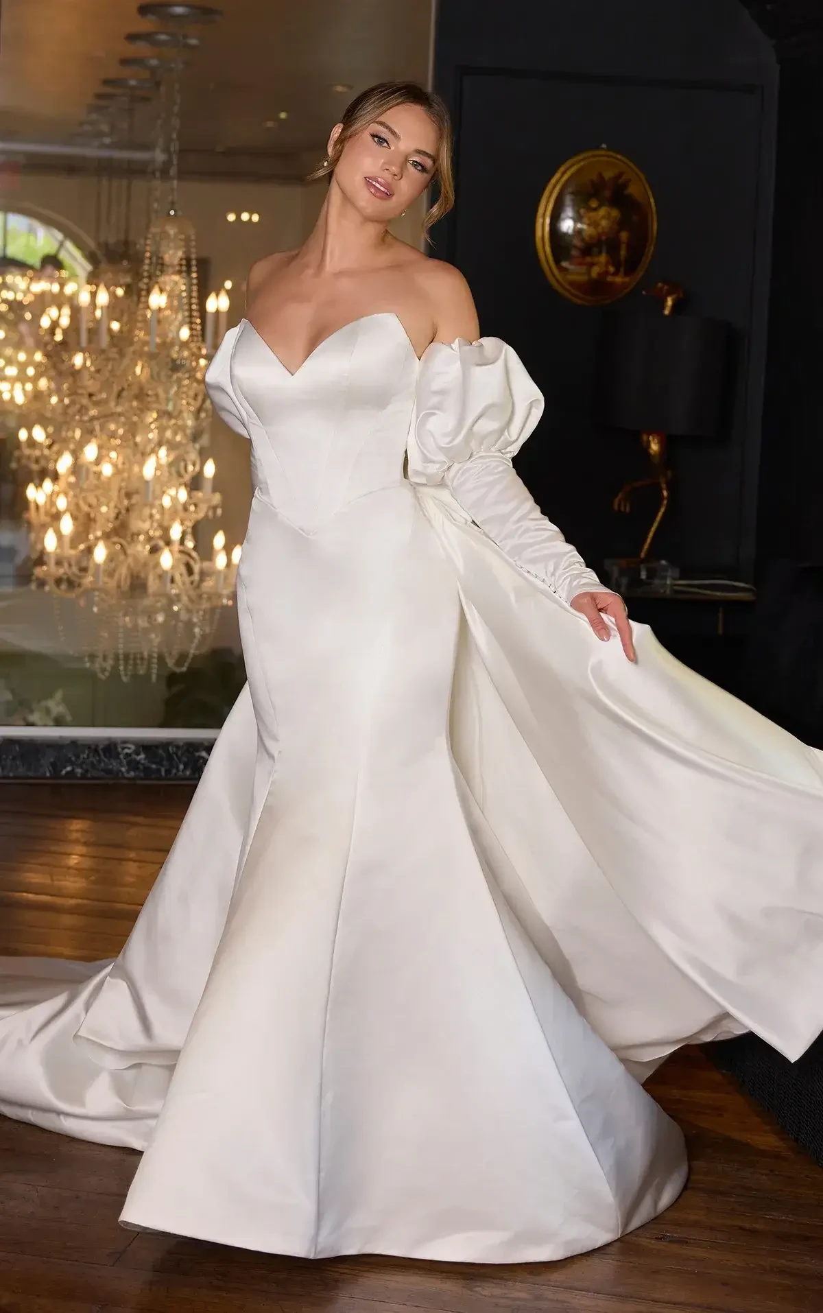 A woman in a stunning white gown with a fitted bodice and dramatic sleeves, posing elegantly in an upscale interior with a chandelier in the background.
