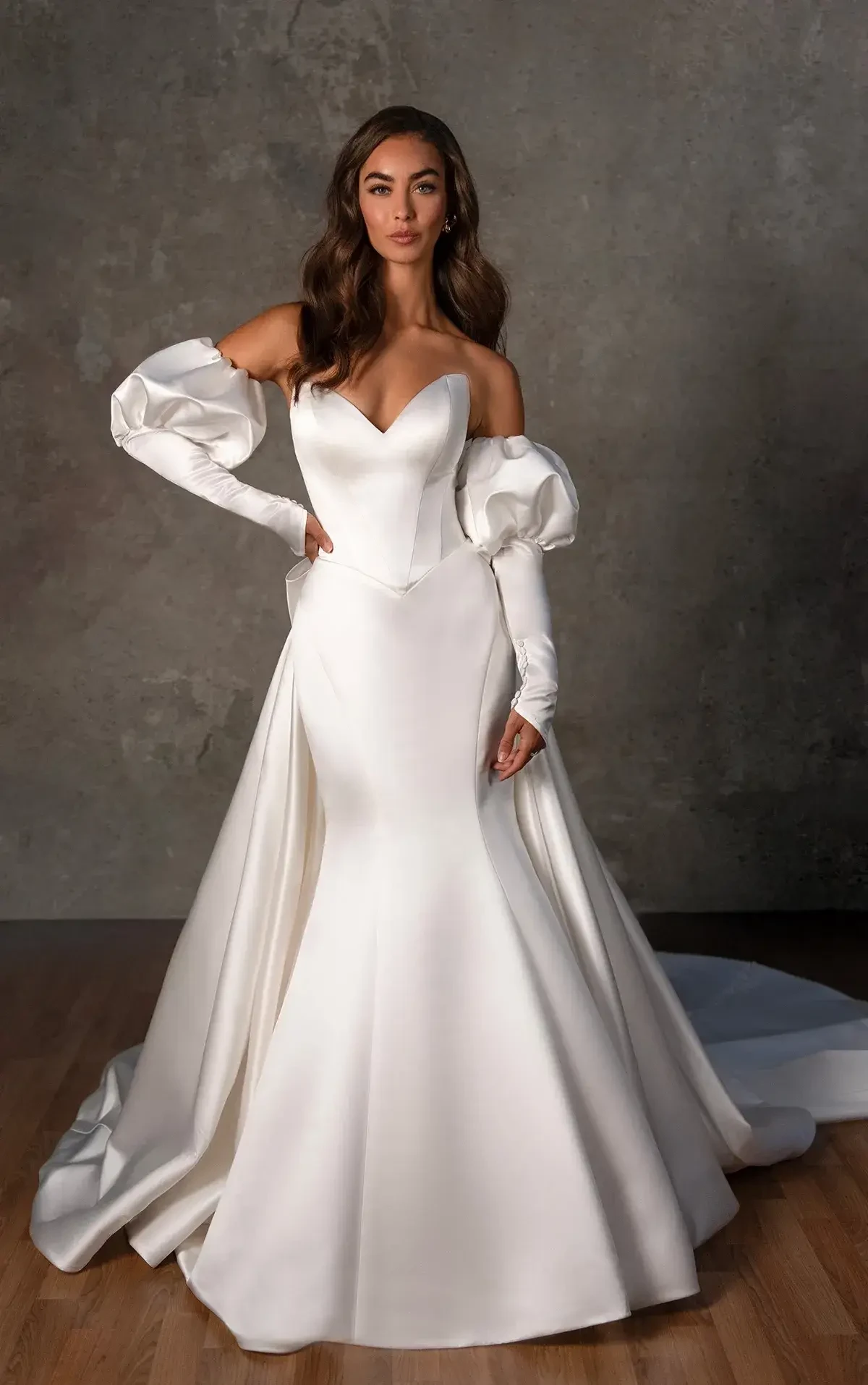 A woman wearing an elegant white wedding gown with off-the-shoulder puff sleeves, standing against a textured gray wall.