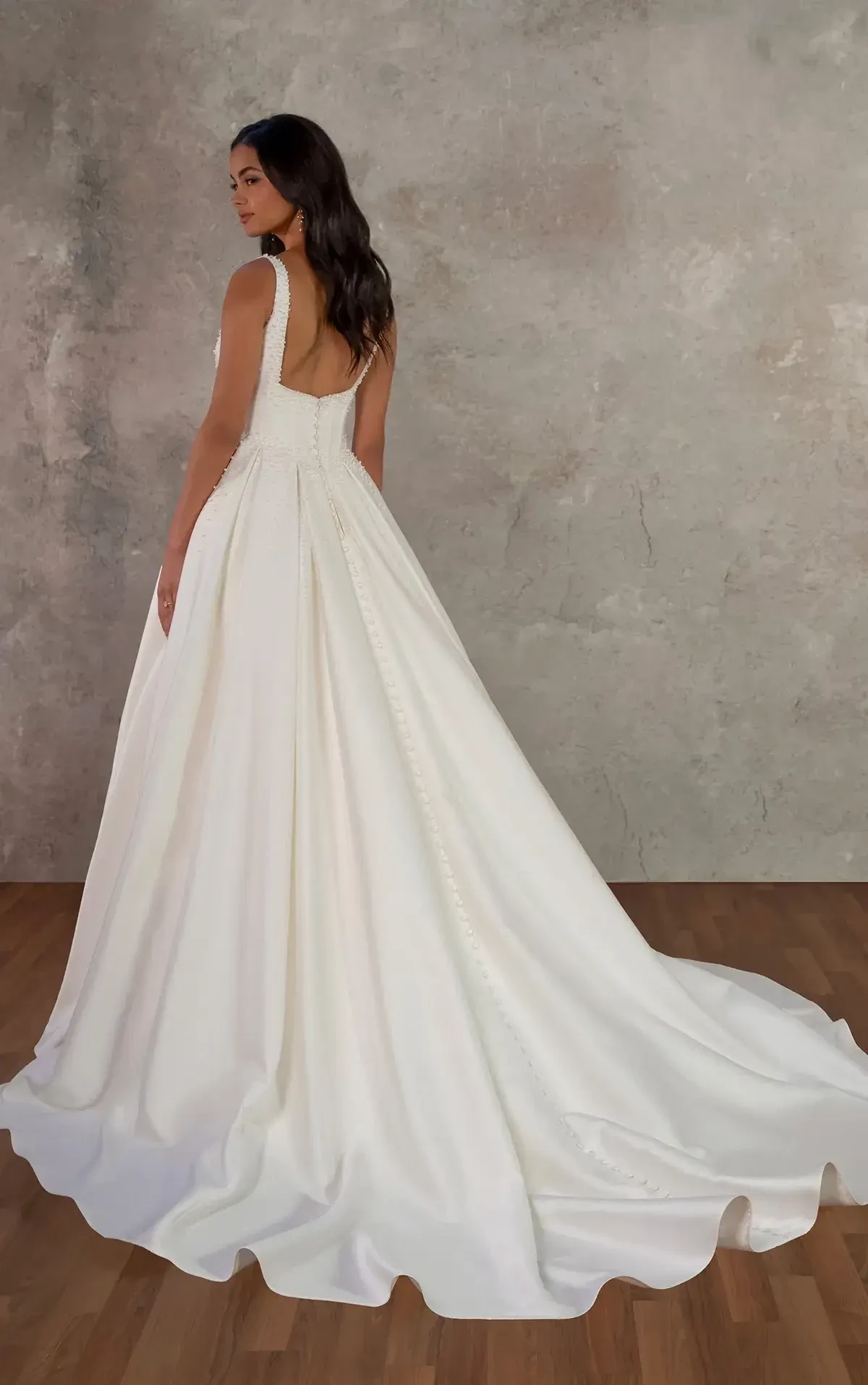 A woman wearing a long, elegant white dress standing with her back to the camera, showcasing the flowing train and neckline. The background is a neutral-toned wall.