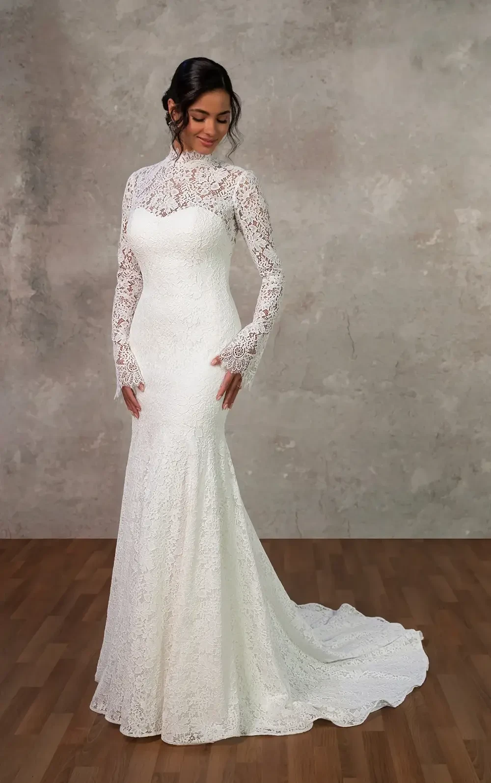 A woman in a long-sleeved, lace wedding gown stands gracefully against a neutral backdrop.