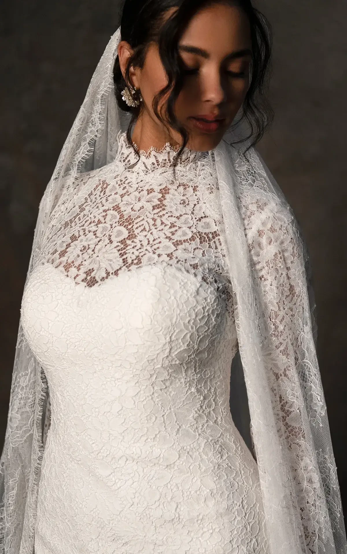A woman in a white lace wedding dress with a high neckline and long veil, looking down.