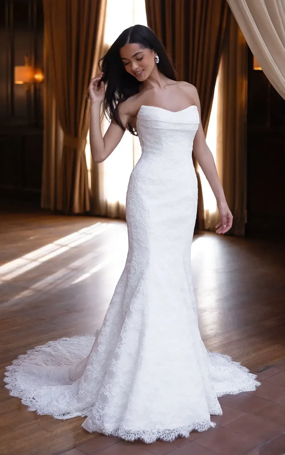 A woman in a strapless white wedding dress stands gracefully, with her hair flowing and a soft smile, in an elegant indoor setting with draped curtains and wooden floors.