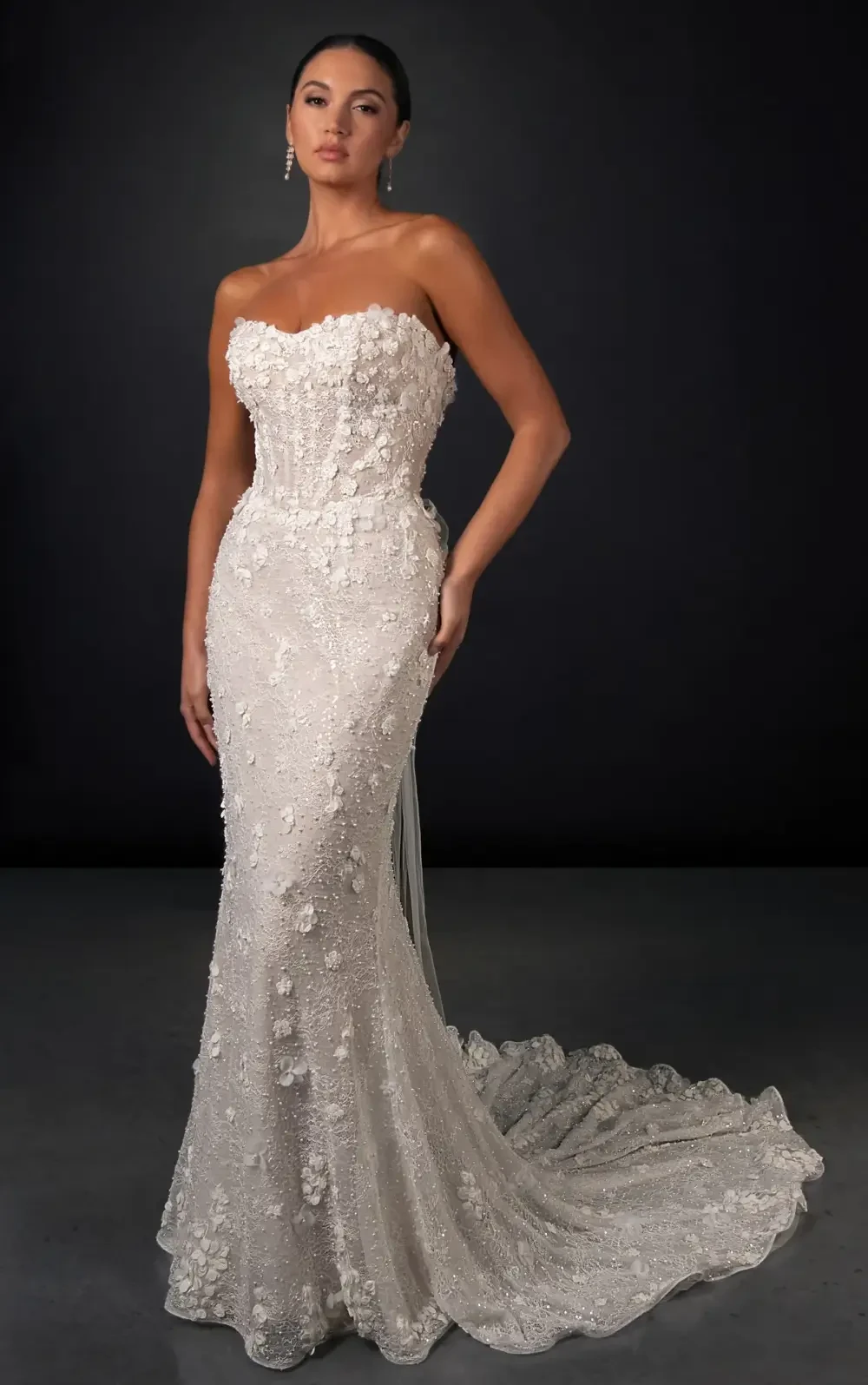A woman in a strapless, fitted wedding gown adorned with intricate floral details and a flowing train, standing against a dark background.