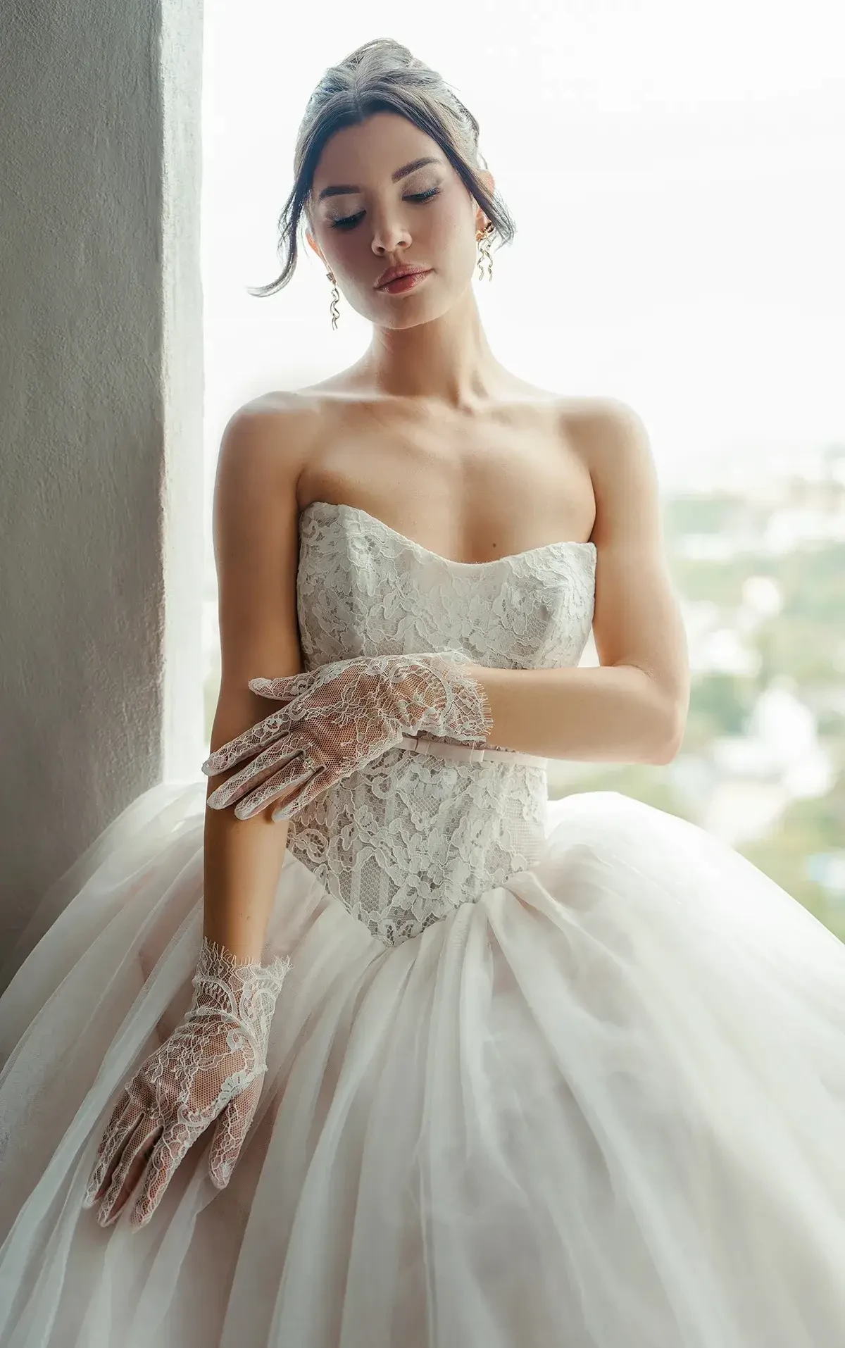 A woman in a wedding dress with lace gloves, standing by a window.