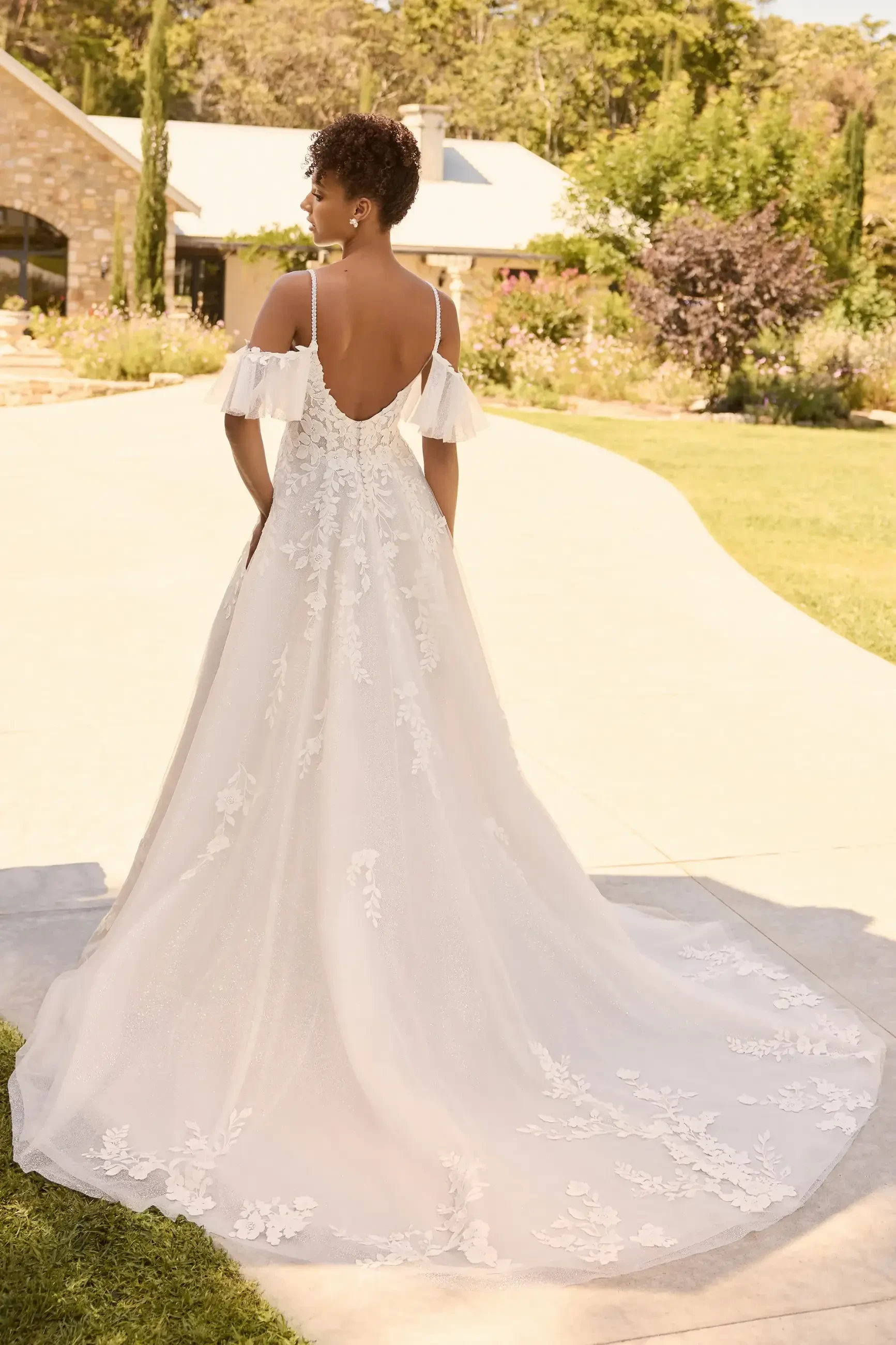 Model is wearing a bohemian A-line gown with floral lace, beaded spaghetti straps, a scoop neckline, layers of glitter and misty tulle, and a chic skirt slit.