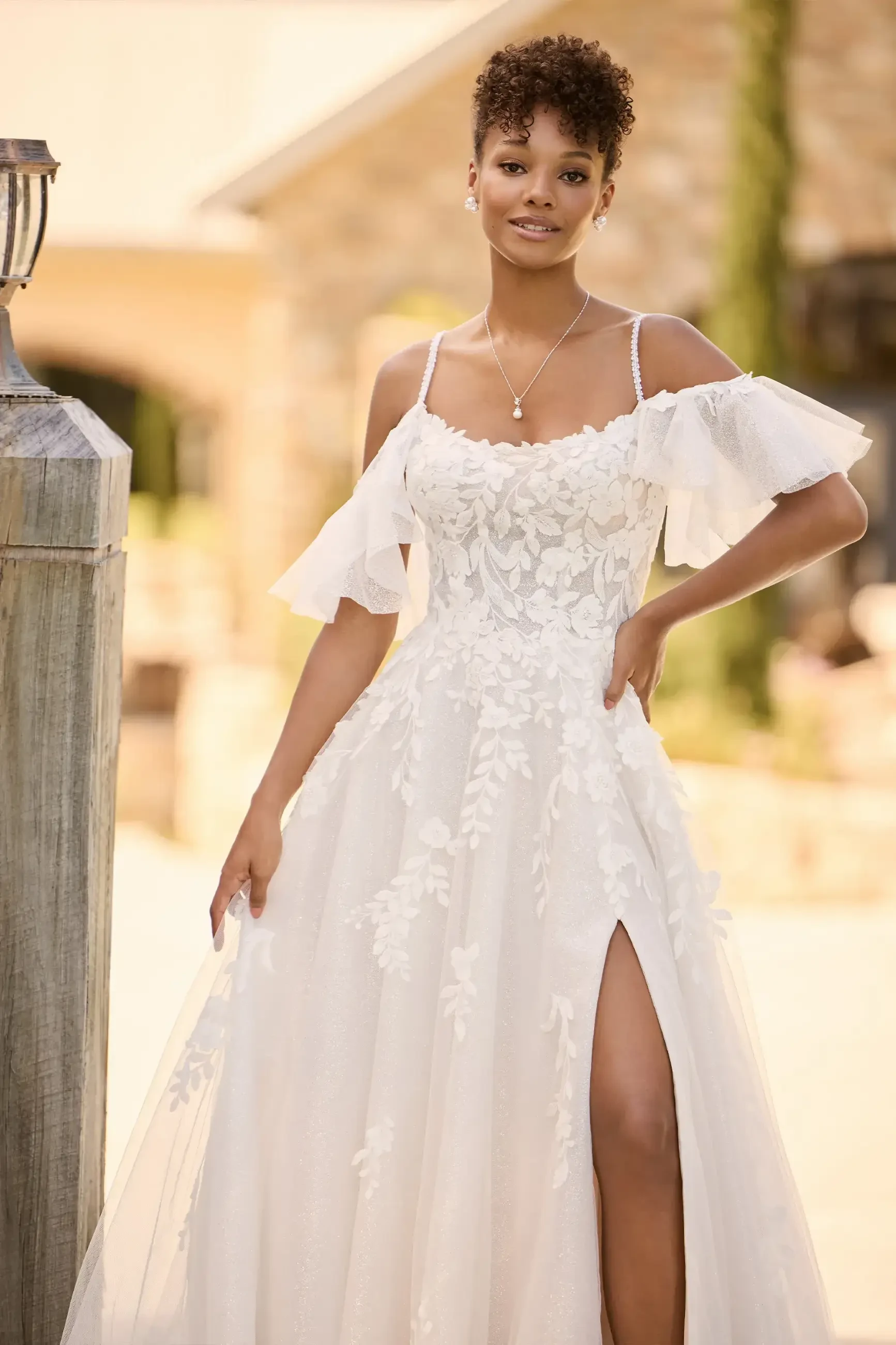 Model is wearing a bohemian A-line gown with floral lace, beaded spaghetti straps, a scoop neckline, layers of glitter and misty tulle, and a chic skirt slit.