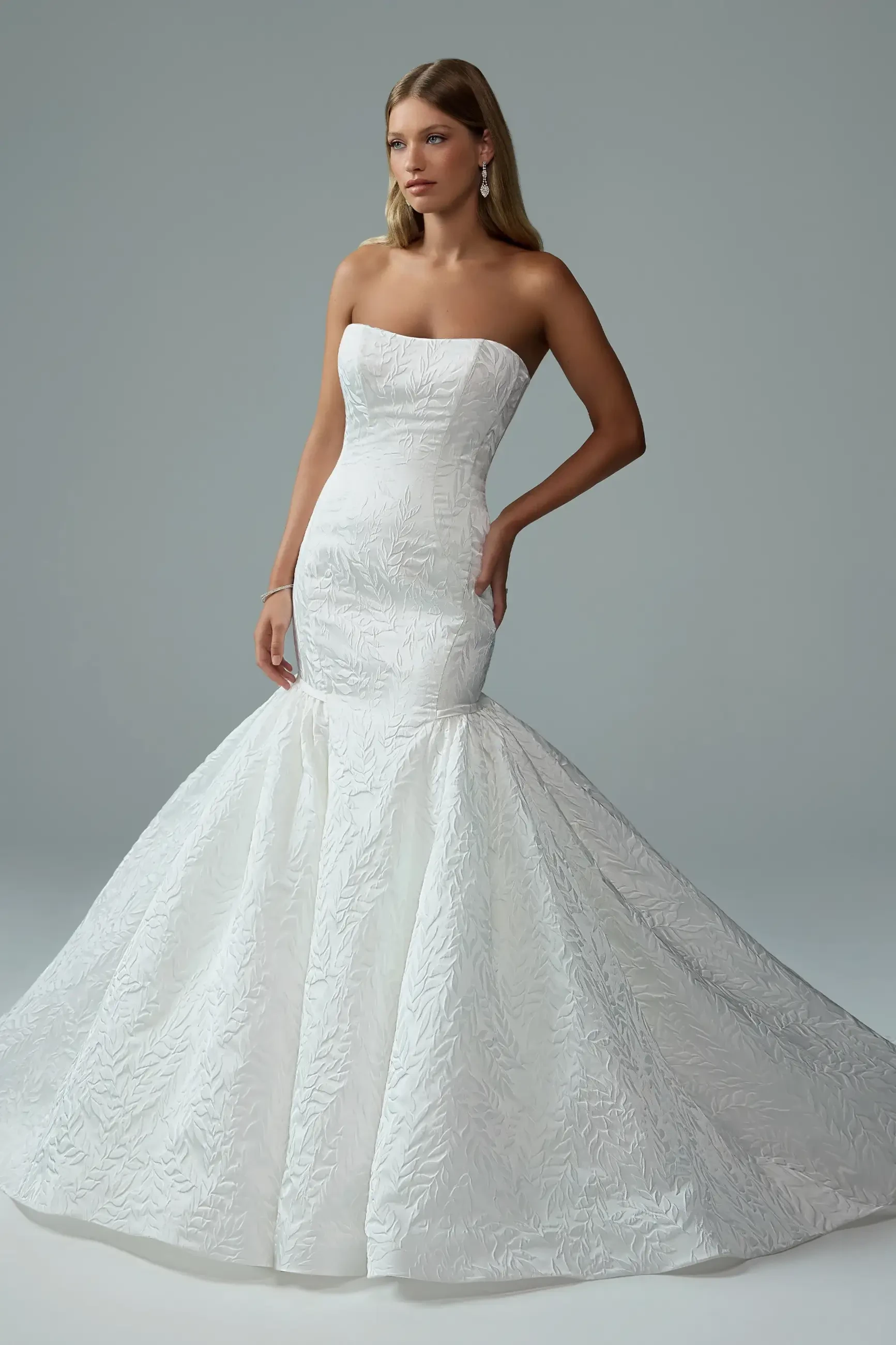 A model posing in a strapless, white mermaid-style gown with intricate detailing and a flared skirt.