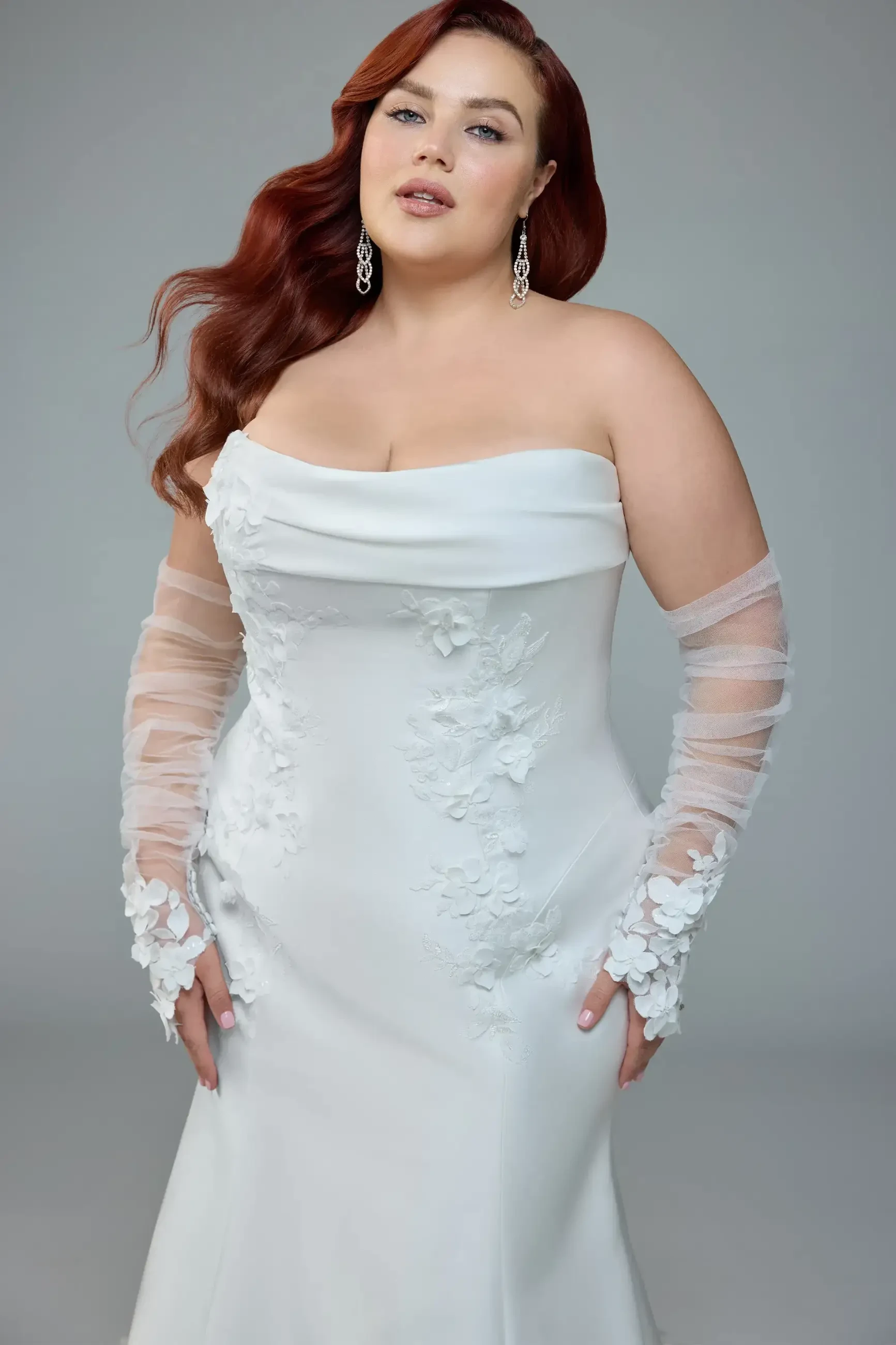 A woman wearing a white off-shoulder gown with floral embellishments and sheer sleeves, styled with long wavy red hair and earrings.